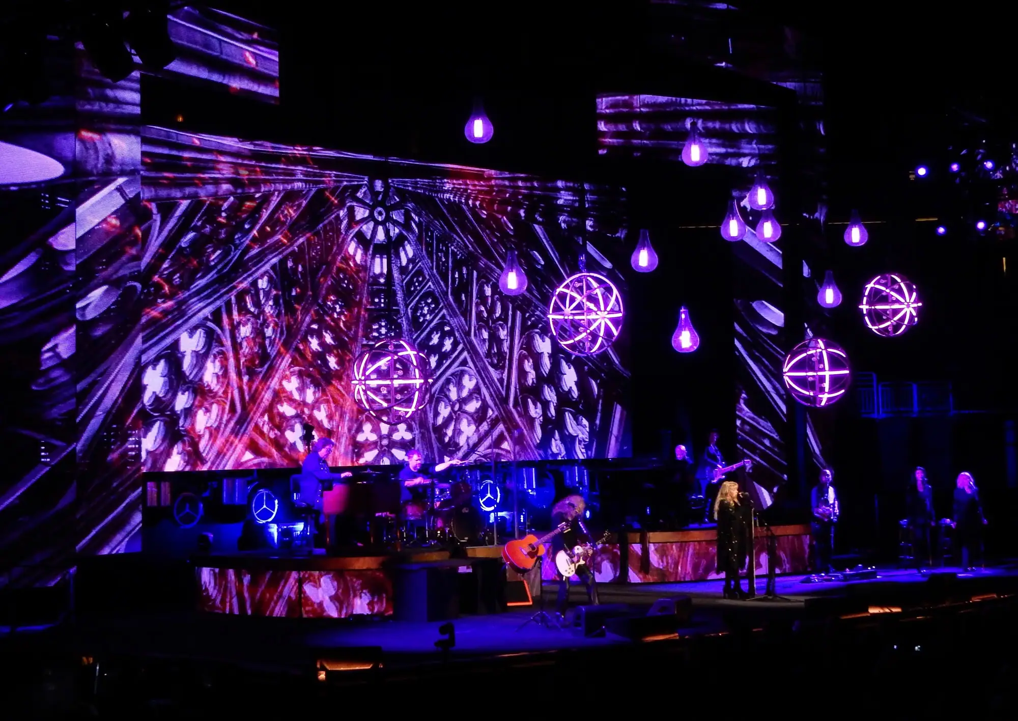 Concierto en un gran escenario con pantallas, esferas de luz y focos morados sobre el grupo