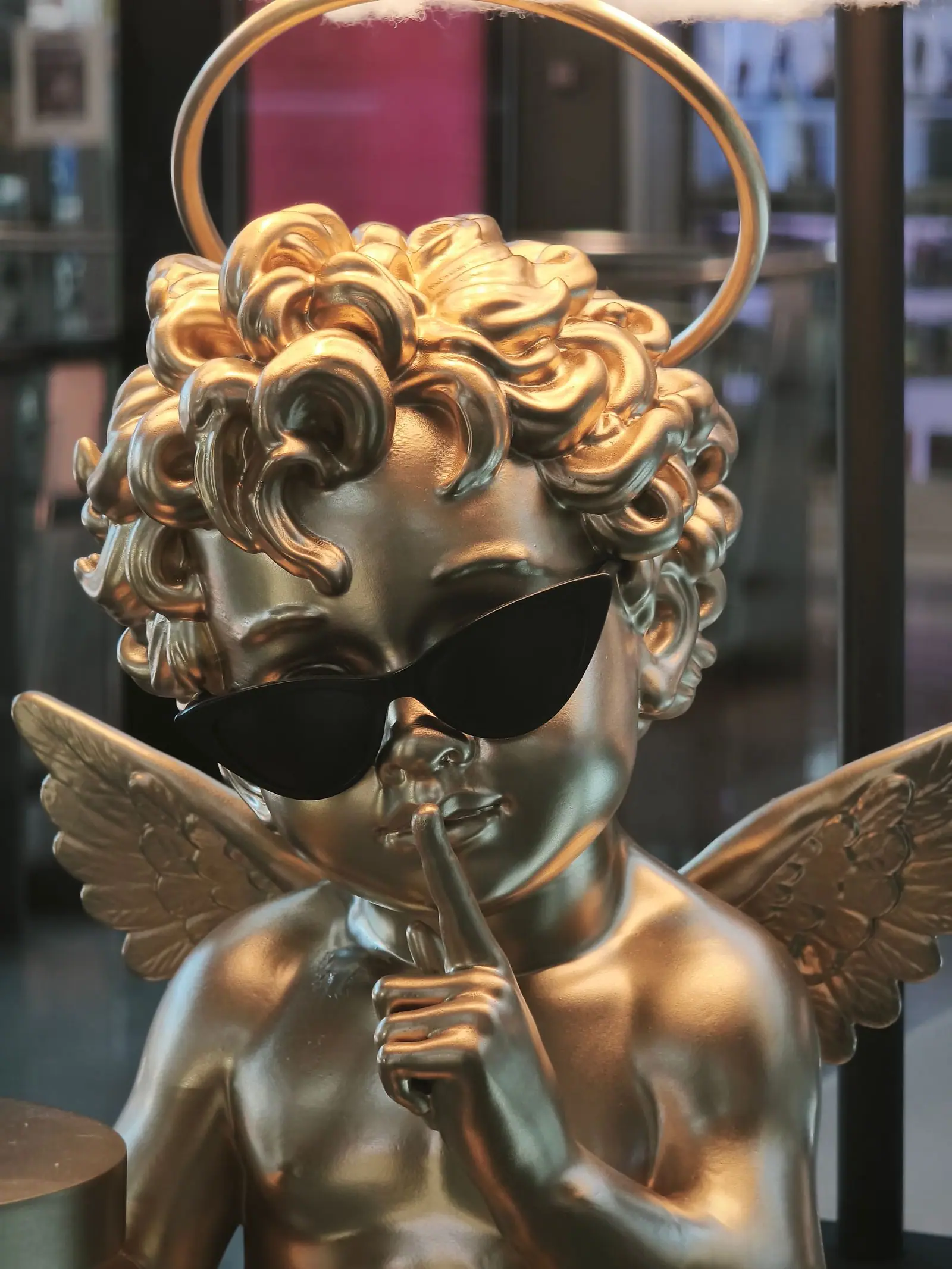 Primer plano del cupido dorado con rizos, aureola y gafas de sol, pidiendo silencio con el dedo