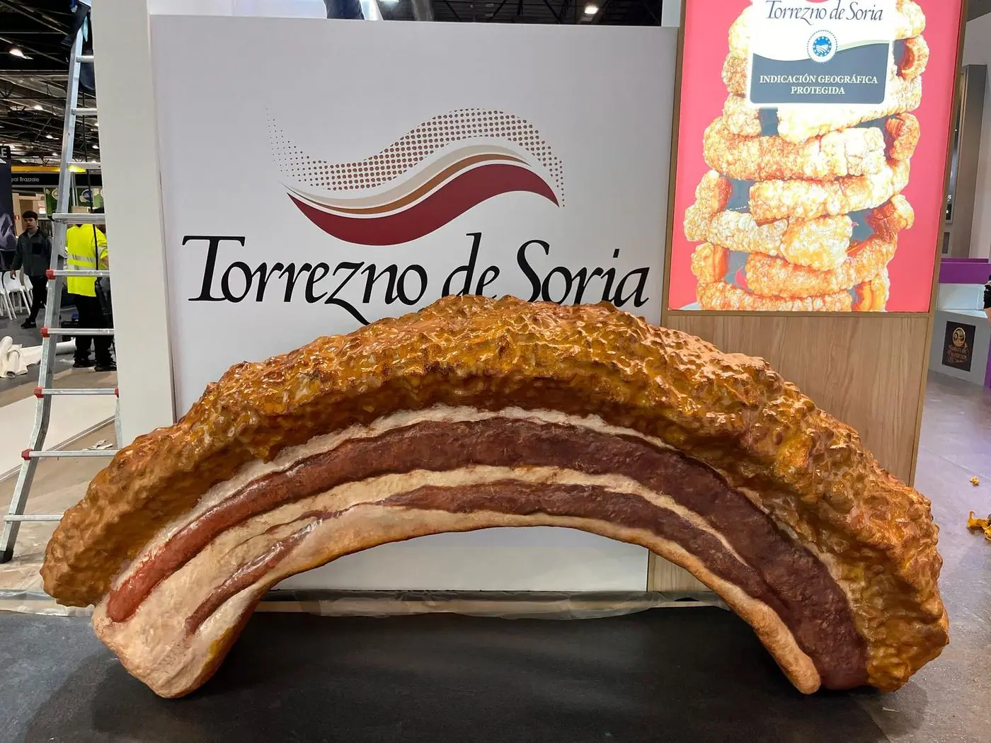 Torrezno de Soria gigante de poliespán, delante de su cartel en una feria