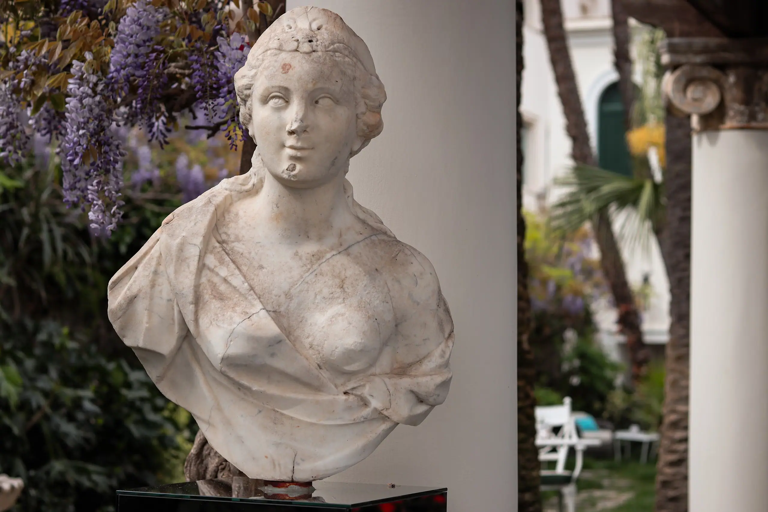 Escultura personalizada de un busto clásico de mujer en piedra blanca, entre columnas y glicinias en flor