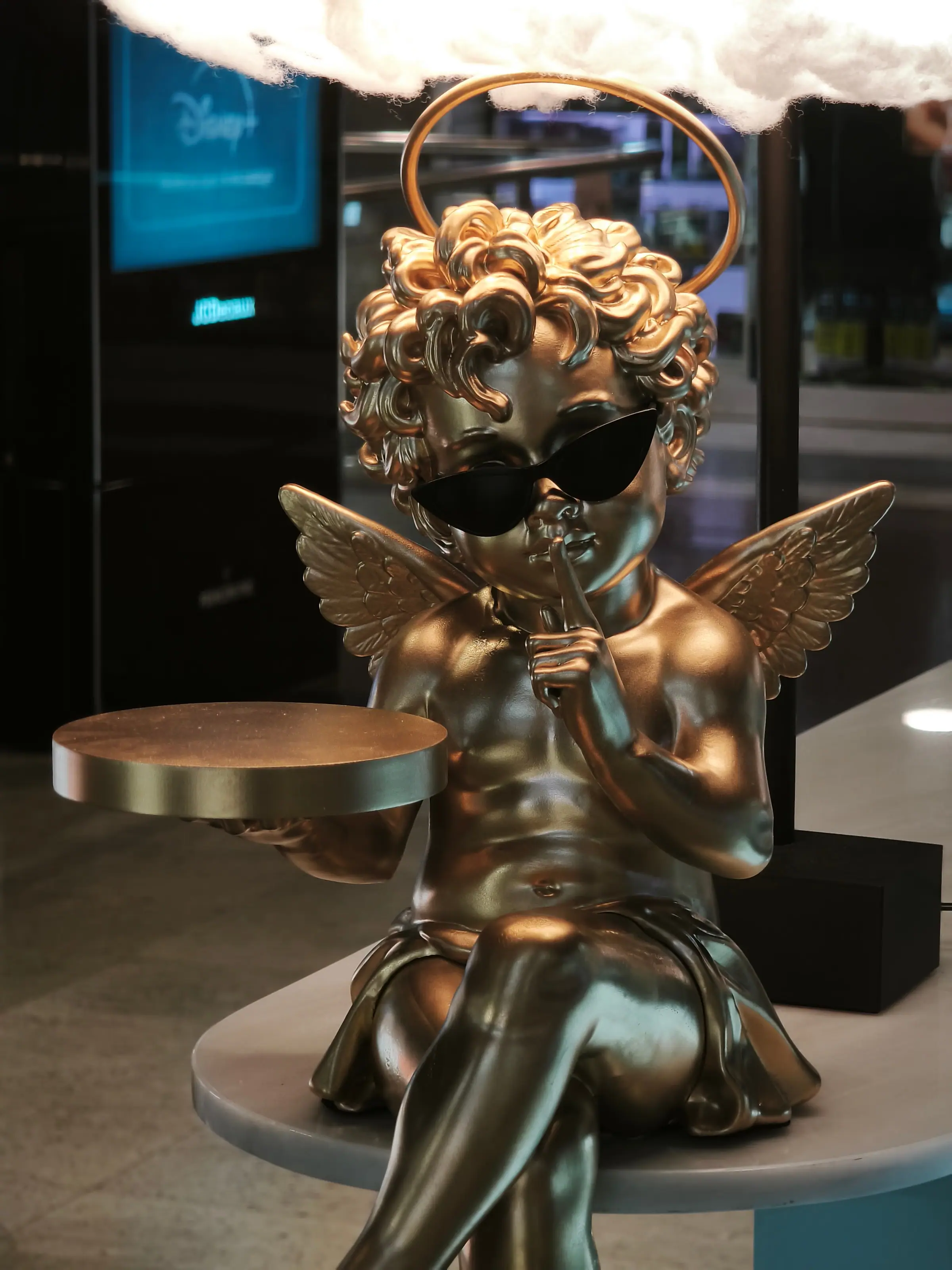 Cupido de resina dorado con gafas de sol sentado bajo una lámpara de nube en un centro comercial