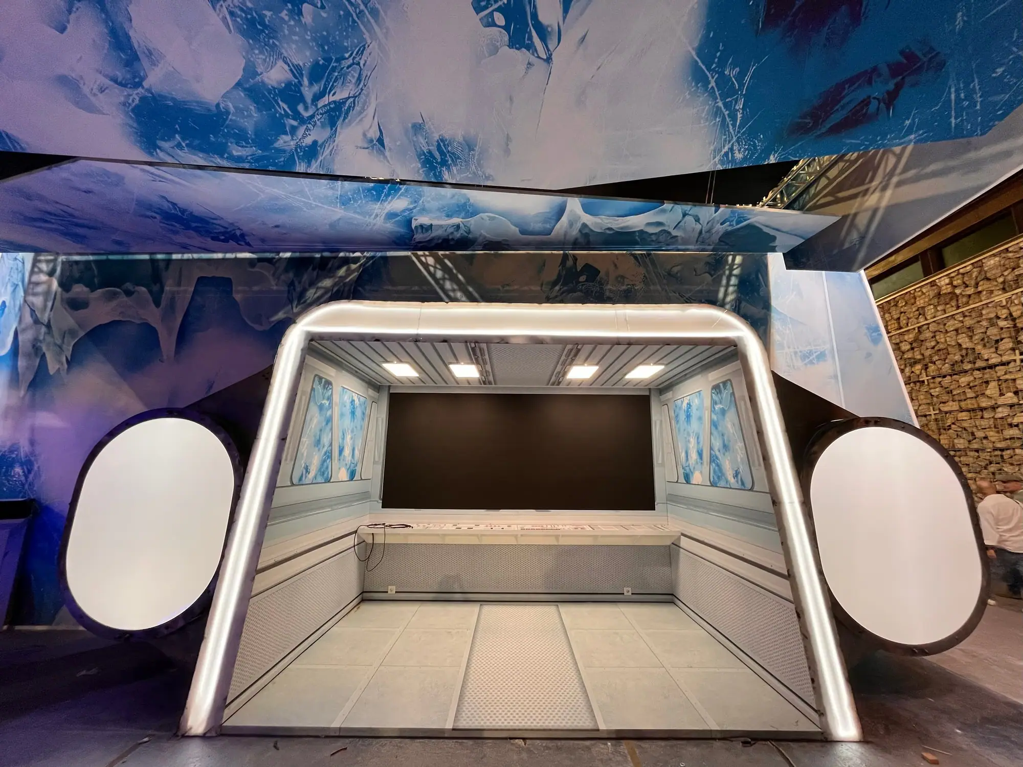 Escenografía de una cápsula futurista con un arco de luz, bancos, una pantalla al fondo y paredes de hielo impresas