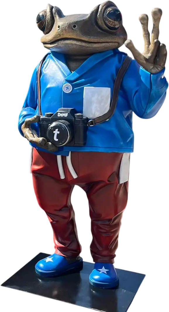 Personaje corpóreo de una rana con cámara de fotos, chaqueta azul y pantalón rojo, sobre su peana