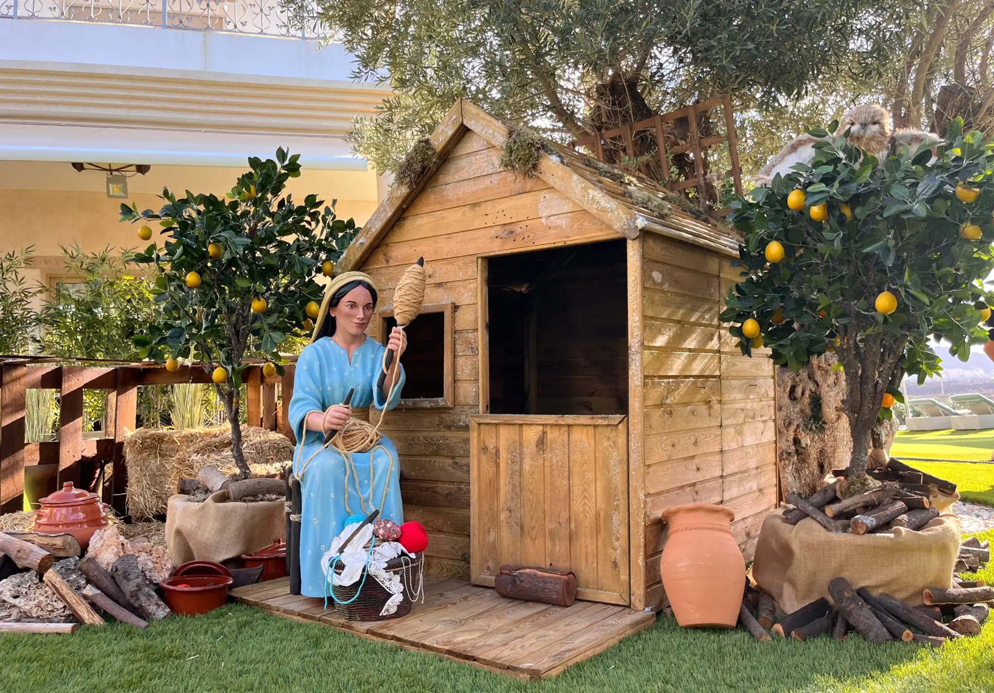 Virgen María a tamaño real hilando a la puerta de una casita de madera en un belén al aire libre