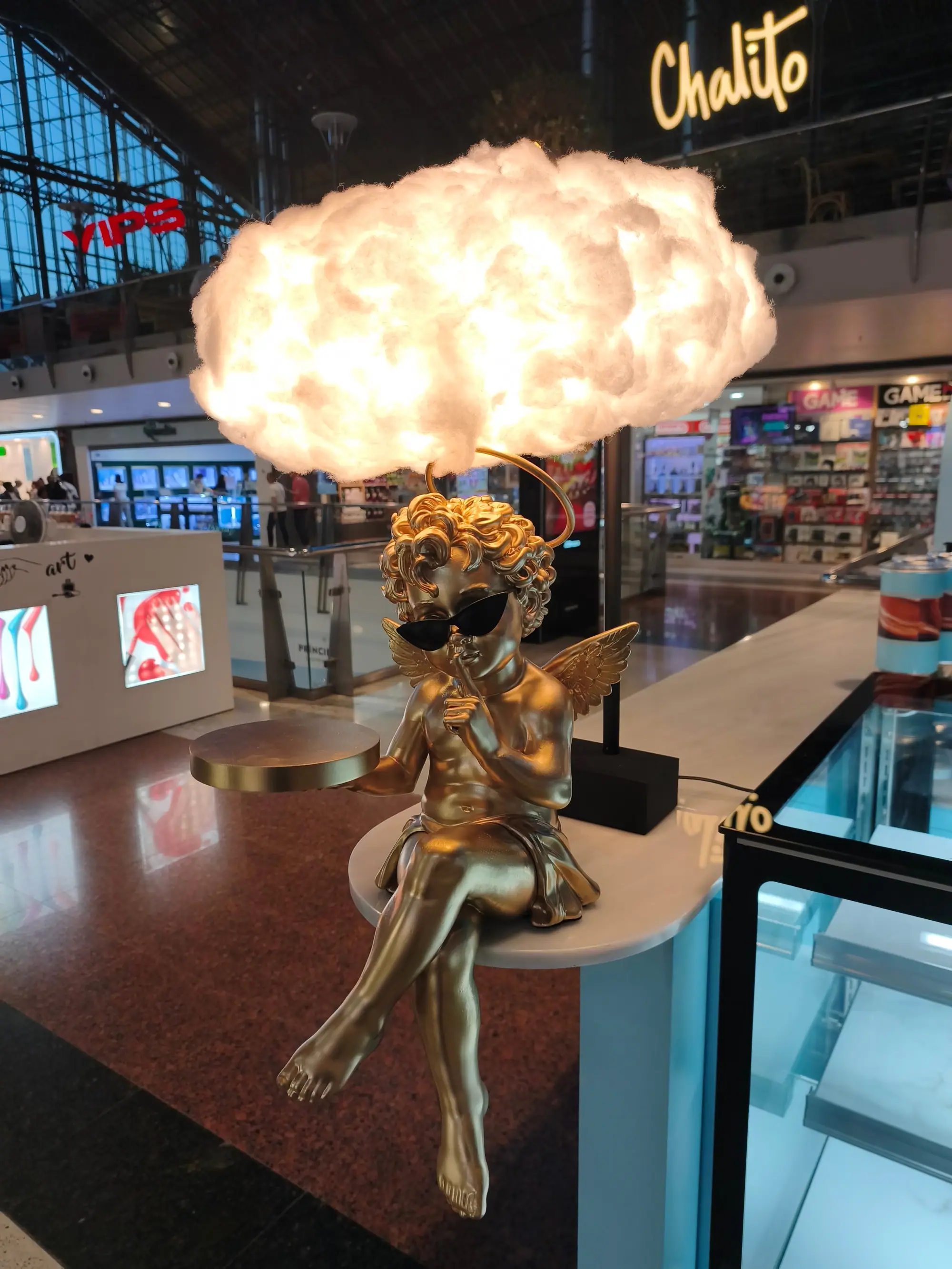Cupido de resina con acabado dorado y gafas de sol, sentado bajo una lámpara de nube en un centro comercial