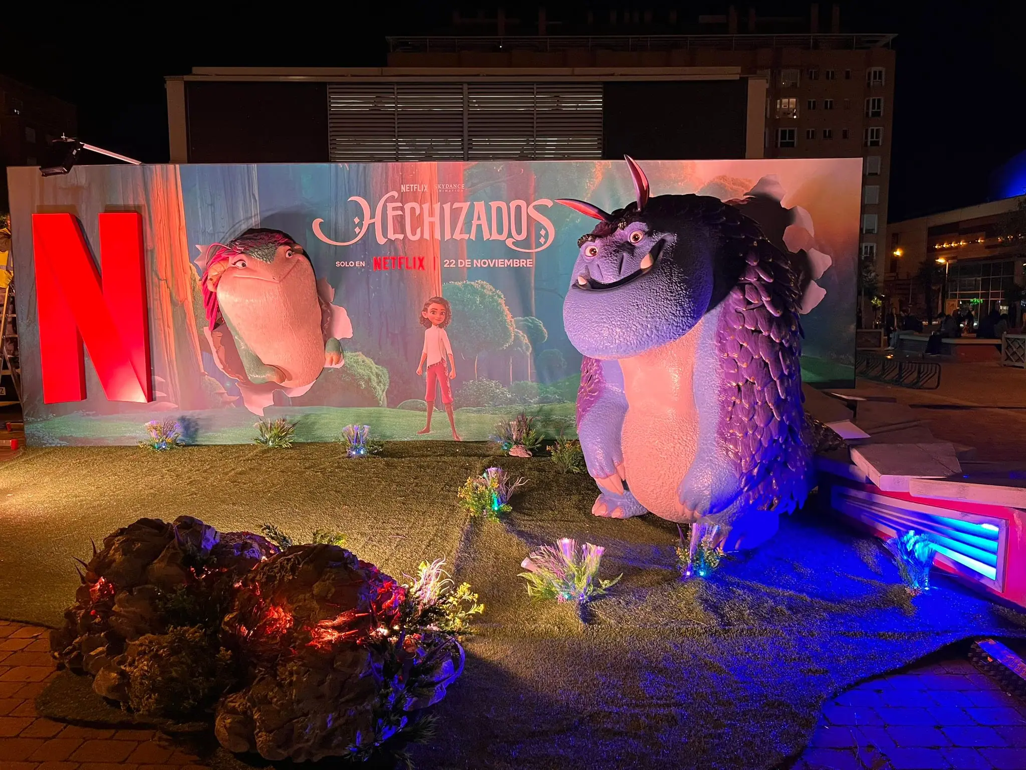 Personaje gigante de la película Hechizados de Netflix, iluminado de noche delante de su cartel en una plaza