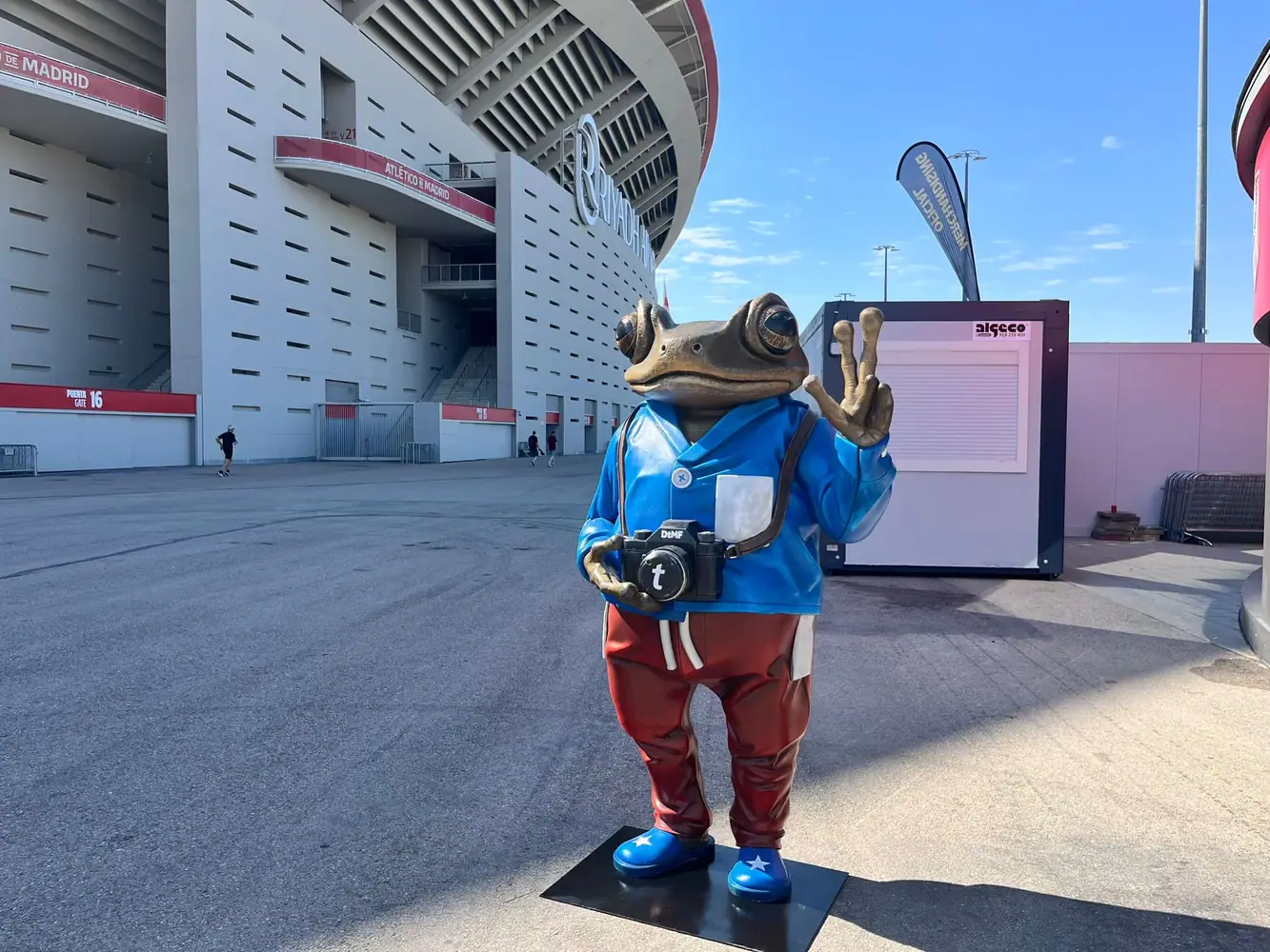Figura de una rana con chaqueta azul, pantalón rojo y cámara de fotos delante de un estadio