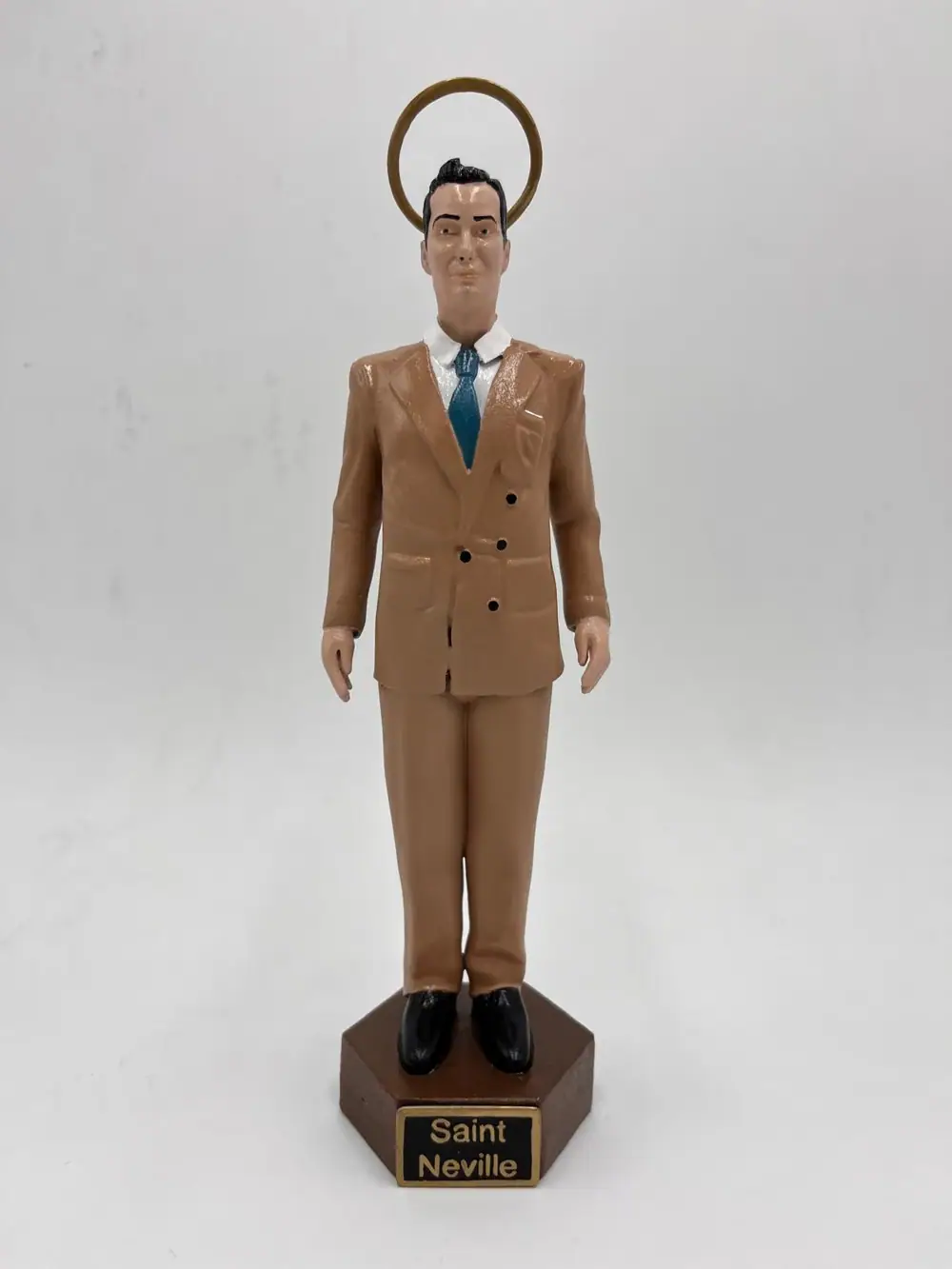 Figura de un hombre con traje marrón y aureola dorada sobre una peana con la placa Saint Neville