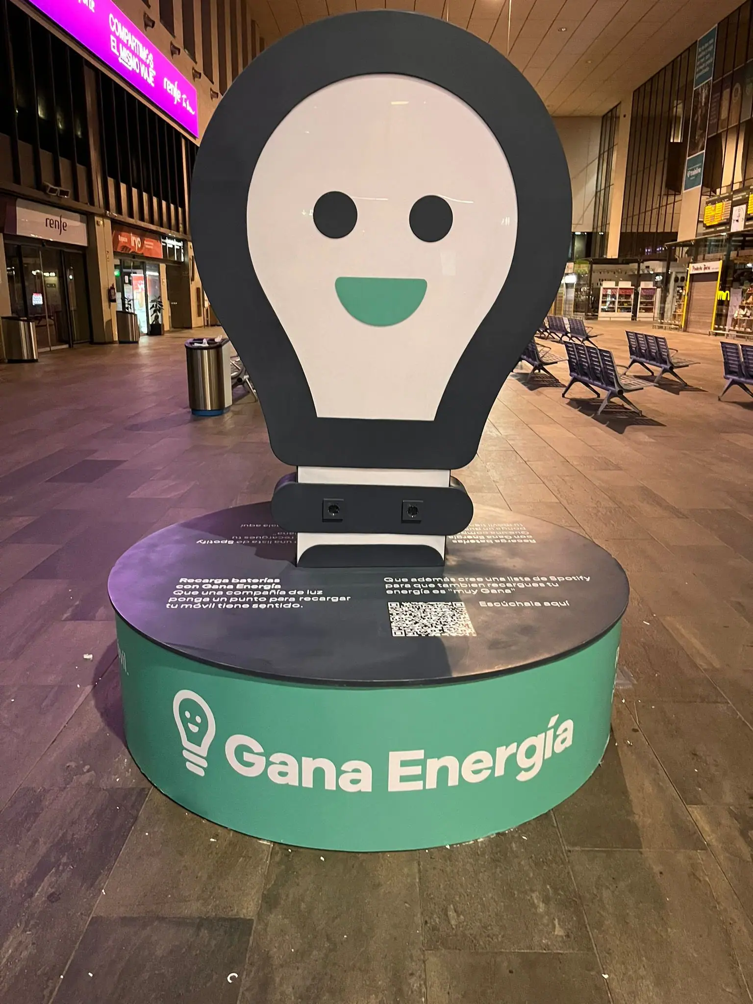 Corpóreo de una bombilla sonriente de Gana Energía con enchufes para cargar el móvil, sobre una peana verde en una estación