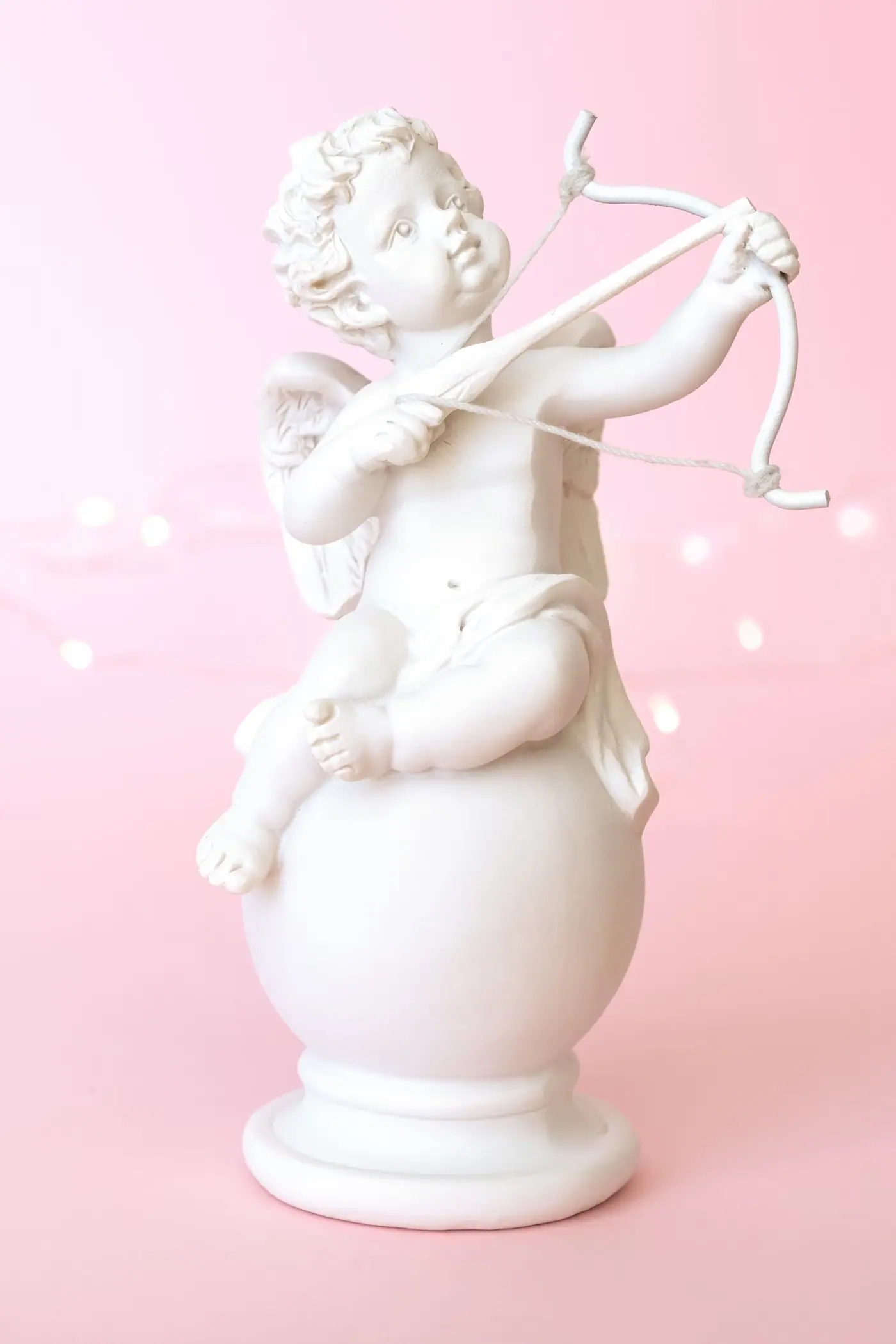 Estatuilla blanca de un cupido con arco y flecha sentado sobre una esfera, con fondo rosa
