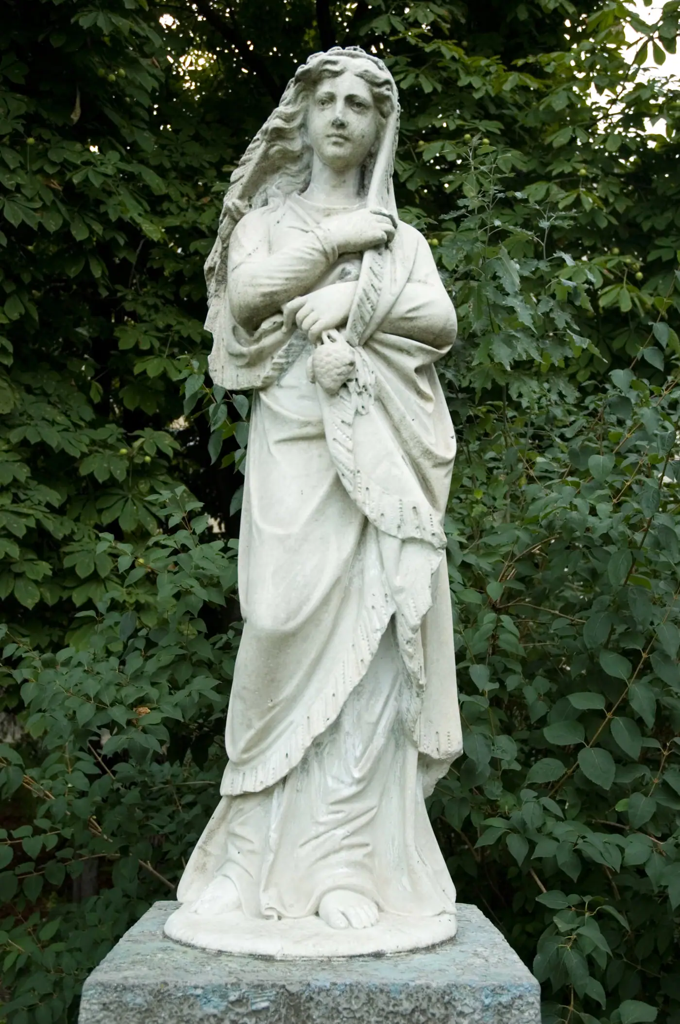 Escultura blanca de una mujer de pie sobre un pedestal, entre los árboles de un jardín