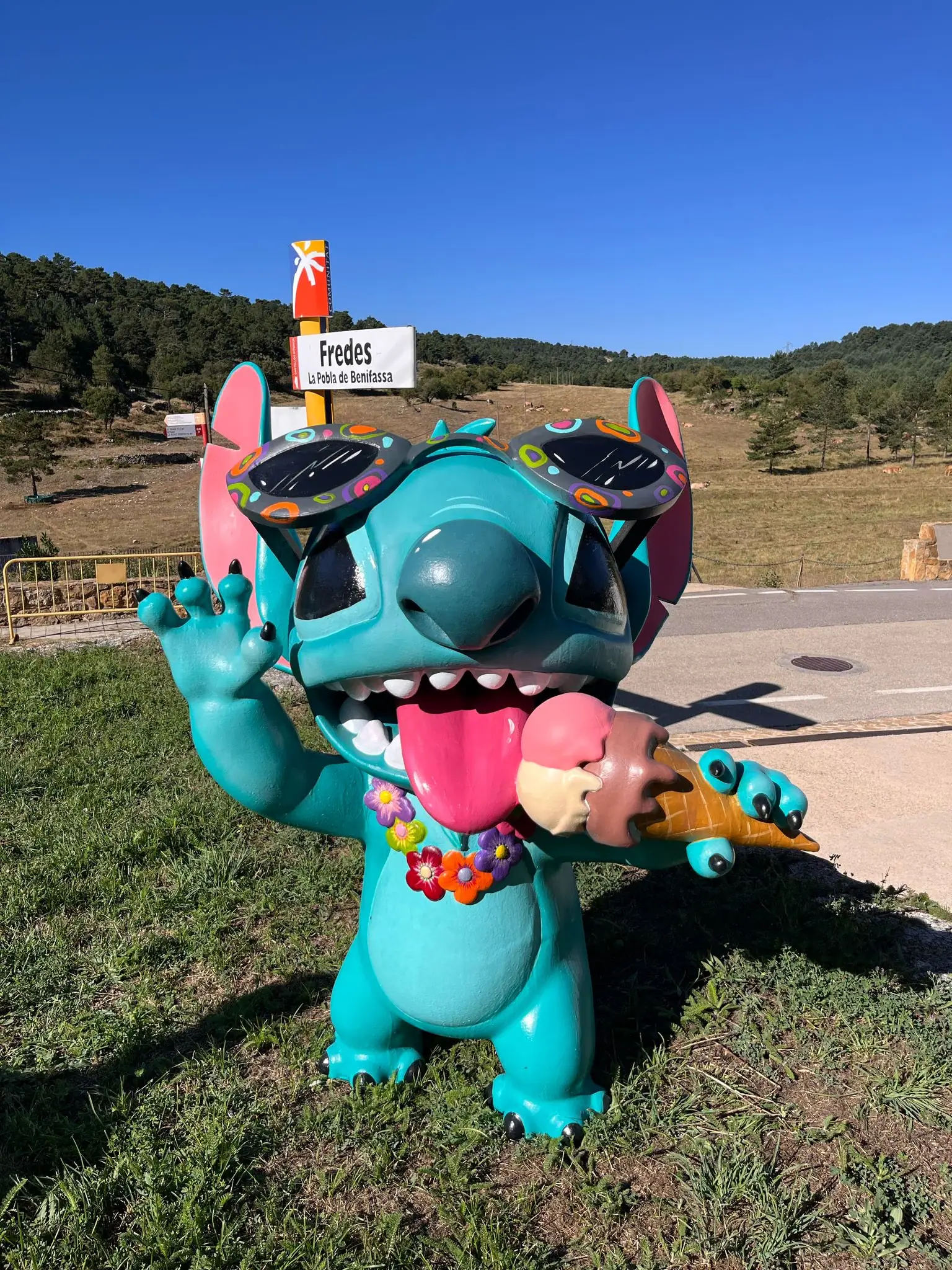 Figura a tamaño real de un personaje azul con gafas de sol y un helado, saludando junto a una carretera de montaña
