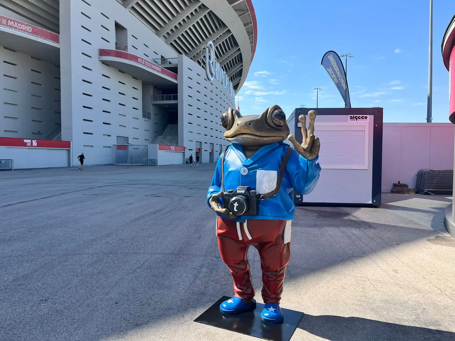 Figura de resina a tamaño real de una rana con chaqueta azul y cámara de fotos, delante de un estadio