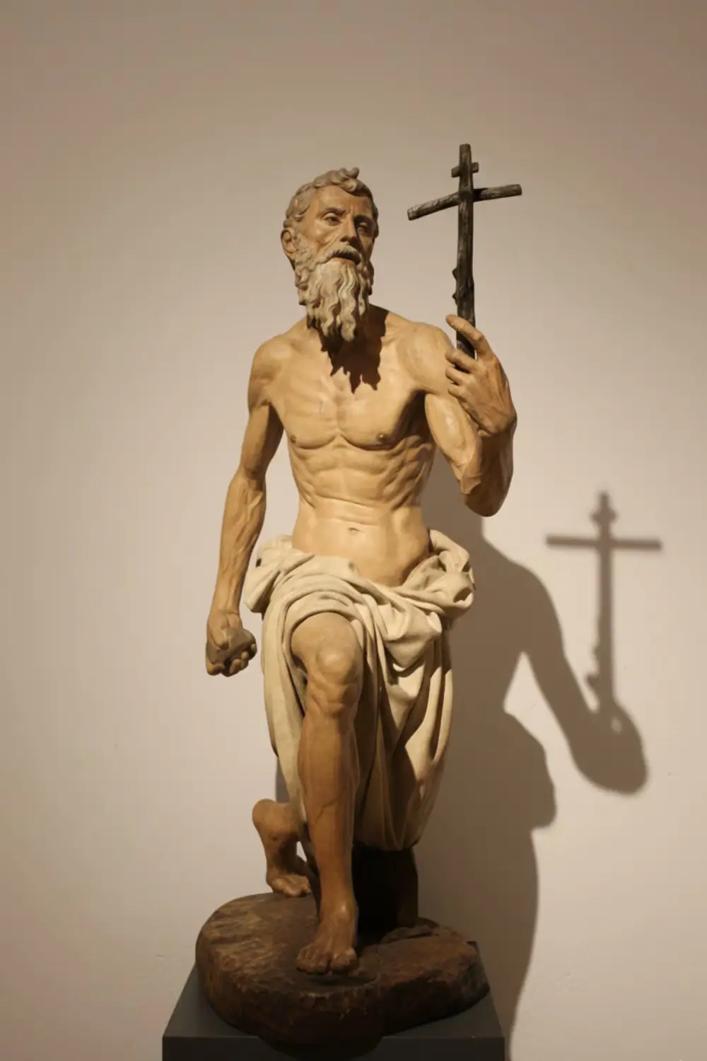 Escultura de un santo penitente arrodillado, con el torso desnudo y una cruz en la mano